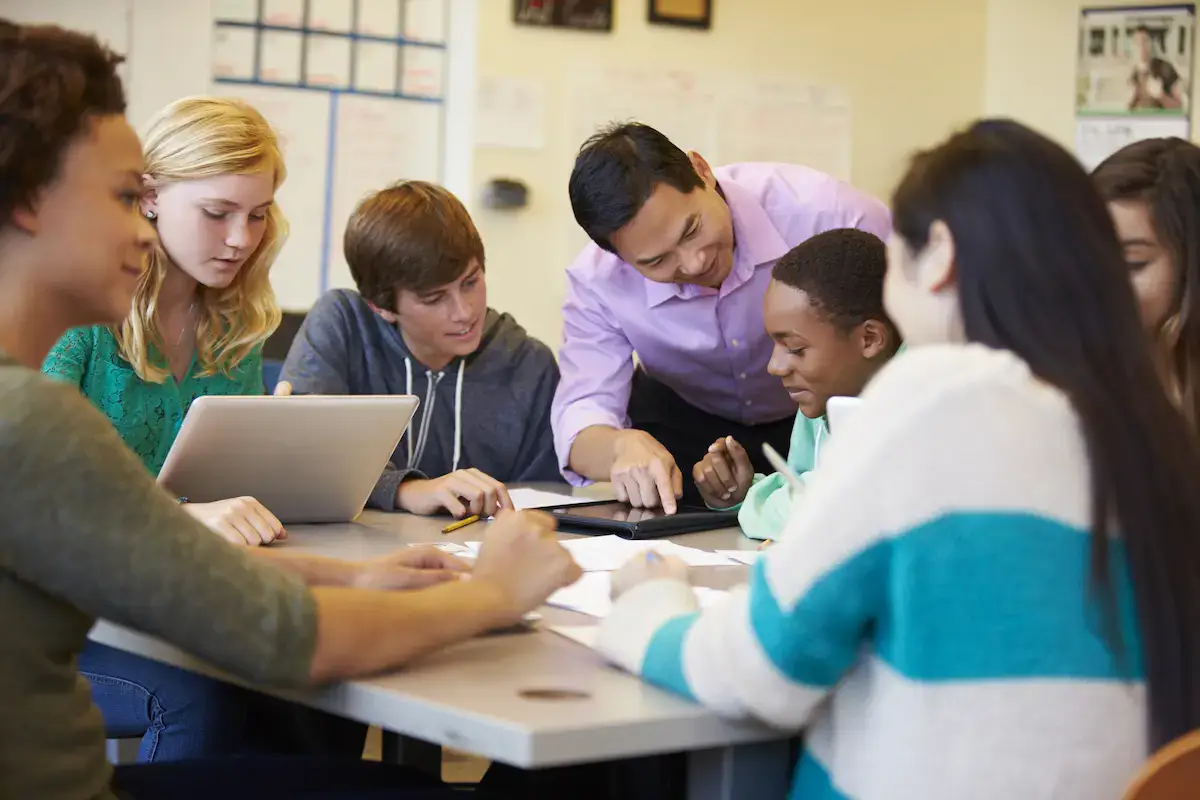 Teacher assisting students with technology and group work in a classroom setting.